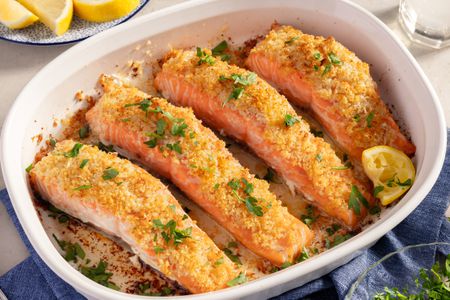 Angled view of a white baking dish of four salmon fillets topped with garlic butter, panko, and parmesan mixture and sprinkled with parsley