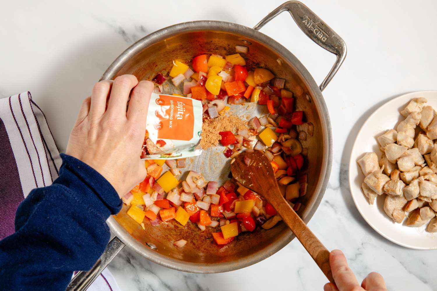 overhead view of hand pouring seasoning mix into pan with veggies, chicken on a plate on the side for One-Pan Chicken Fajita Pasta
