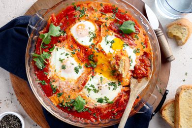 Microwave Shakshuka in a Circular Pyrex Dish on a Circular Tray Surrounded by Slices of Bread on the Counter, a Small Bowl of Cracked Pepper, and a Glass of Water