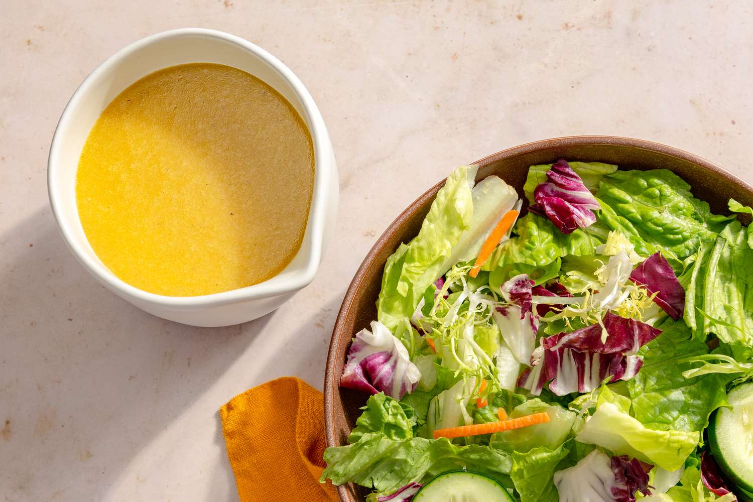 Overhead view of a white bowl of Miso-Maple Dressing next to a bowl of lettuce and cucumbers all on a tan countertop