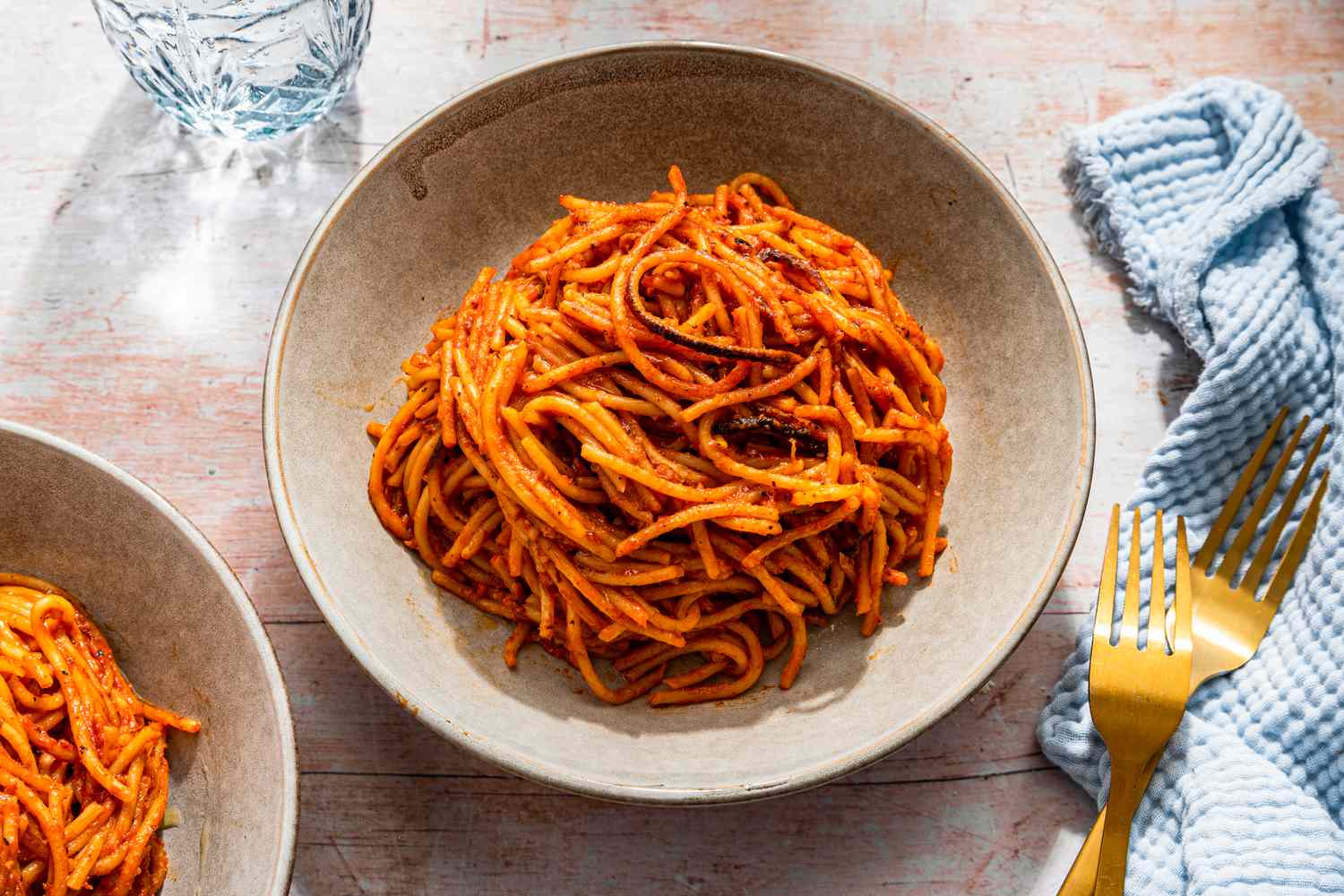Bowlful of assassin's spaghetti at a table setting with more in a bowl, a glass of water, utensils, and light blue kitchen towel