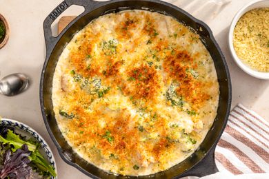 Skillet chicken divan in a cast iron skillet at a table setting with a bowl of salad, a bowl of parmesan, a small bowl of chopped parsley, and a utensils
