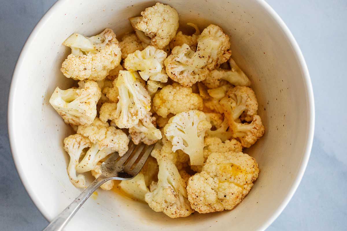 Cauliflower florets in a mixing bowl and coated in spices. A fork is inside the bowl with the cauliflower.