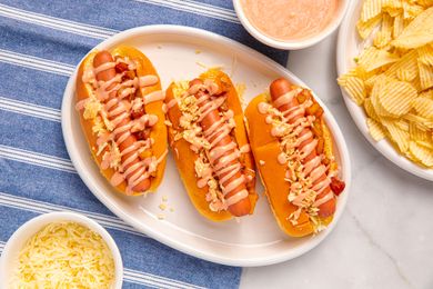 Three hot dogs on a plate with toppings and sauces accompanied by a side of chips and shredded cheese