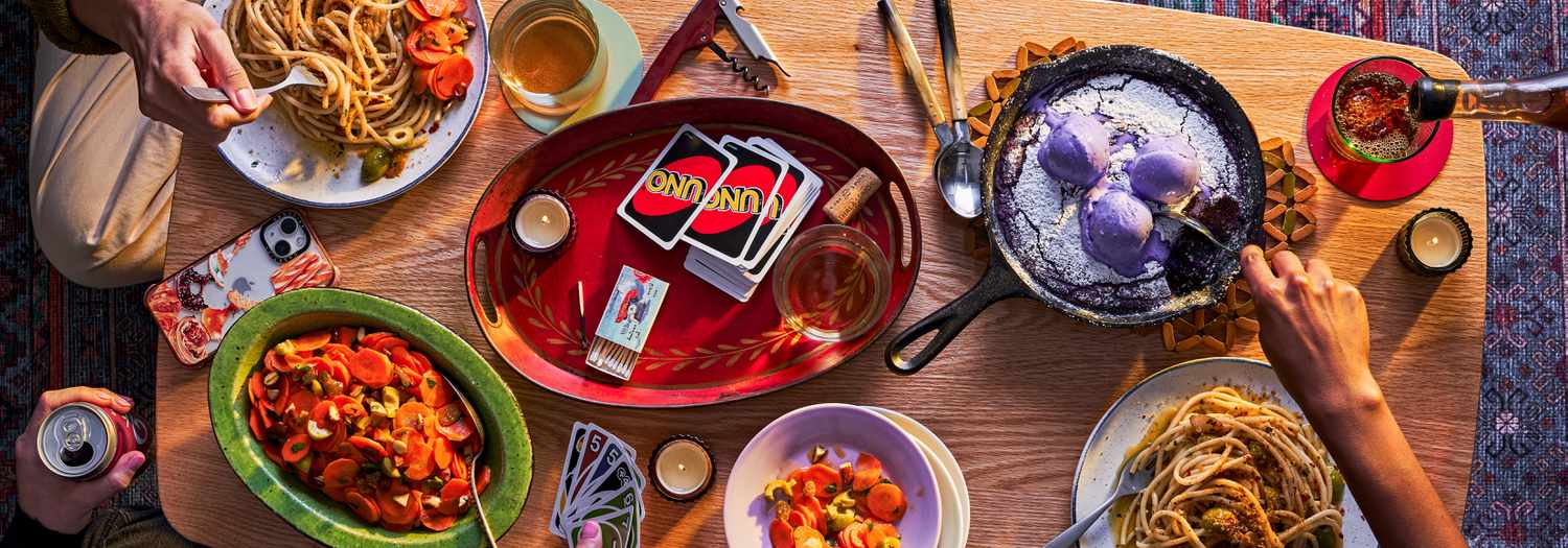 Coffee table filled with food including pasta, carrot salad, and ube skillet cookie, Hands are digging into the food and playing uno