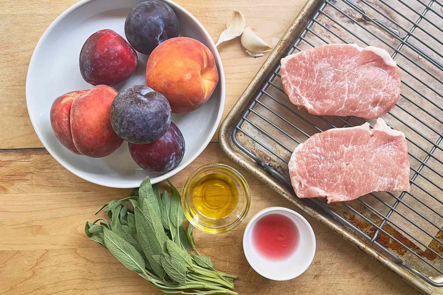 Pork chops on a tray peaches and plums on a plate fresh sage and small bowls of olive oil and vinegar placed on a wooden surface