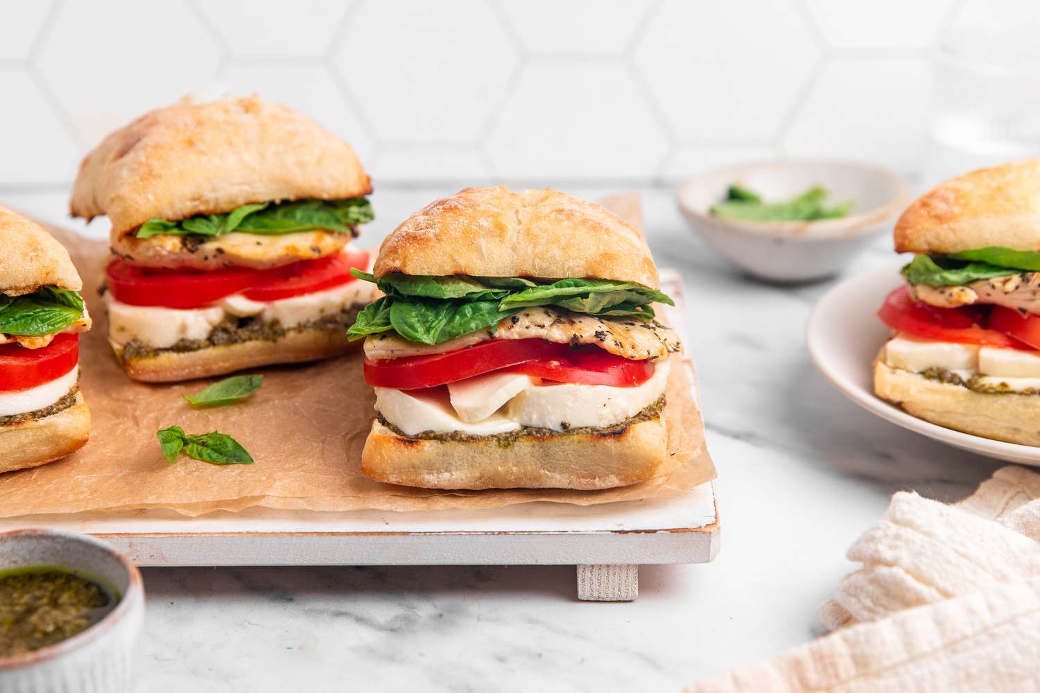 Grilled Chicken Caprese Sandwiches on a Parchment Lined Tray, and on the Counter Next to the Tray, Plates With Individual Ingredients, a Bowl With More Pesto, a Plate With Another Sandwich, ad a White Kitchen Towel