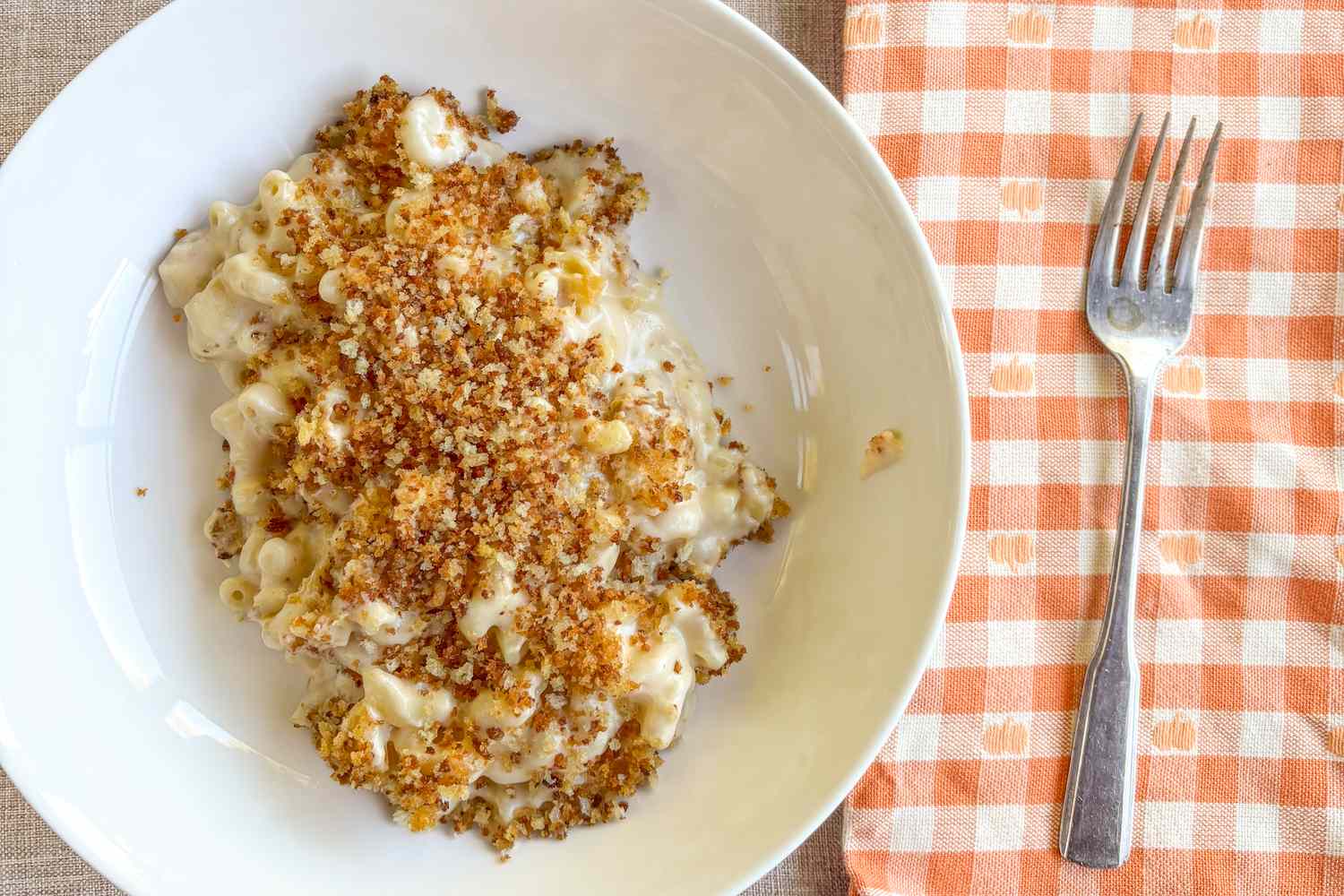 Plate of creamy macaroni and cheese topped with breadcrumbs beside a fork on a checkered napkin