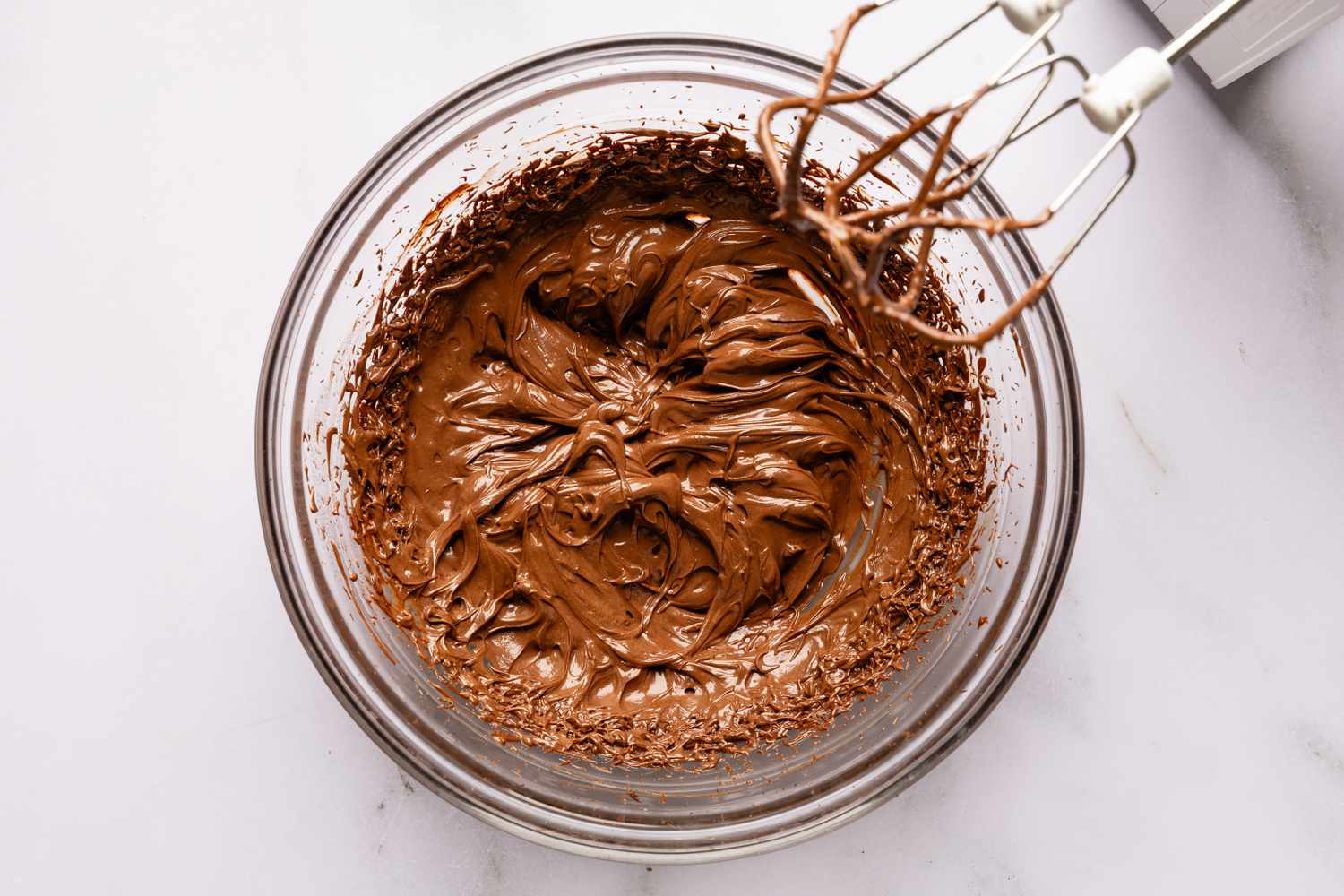 Overhead view of a clear glass bowl and hand mixer after beating the cream and chocolate mixture with the mixer for Easy Brooklyn Blackout Cake recipe