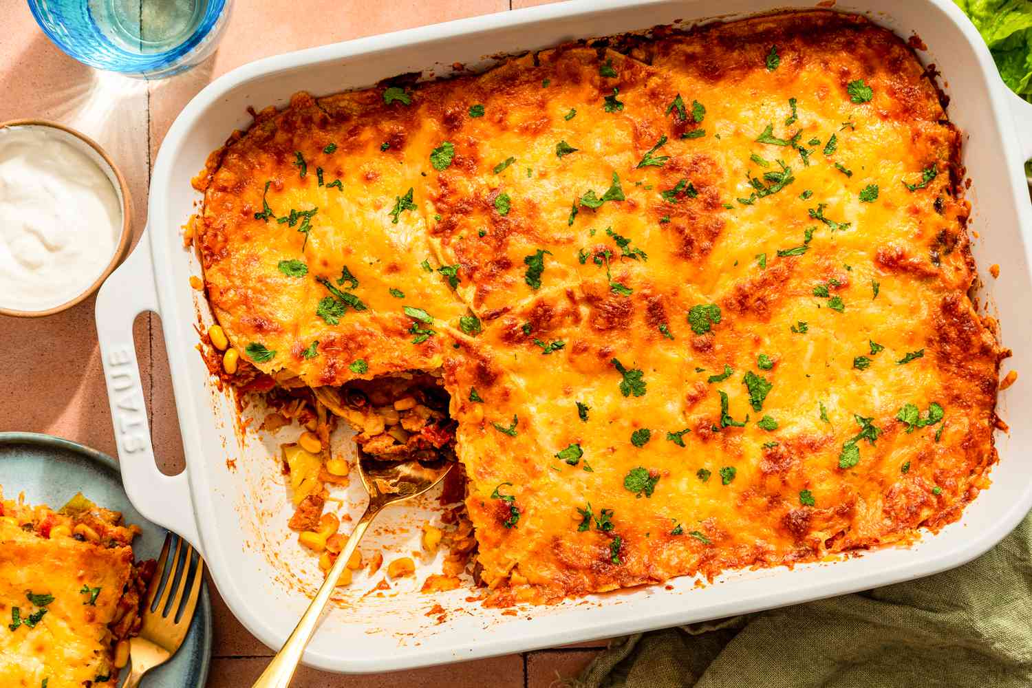 Chicken taco casserole (with a serving cut out) at a table setting with glasses, a serving on a plate, a side salad, a bowl of sour cream, and a kitchen towel