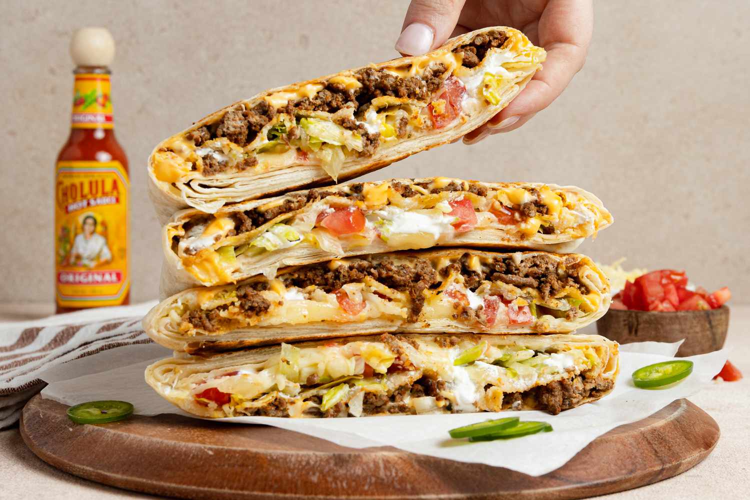 Hand Removing a Wrap from a Stack of Copycat Crunchwrap Supreme Wraps Cut in Half on Parchment Paper Over a Wooden Board