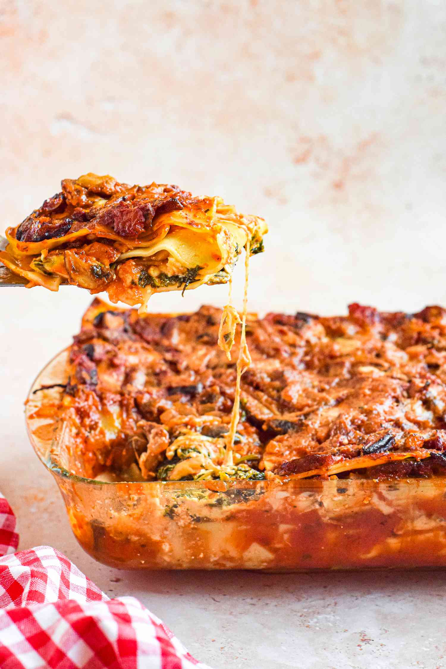 Slice of Vegetarian Spinach and Mushroom Lasagna Lifted from Casserole Dish with More Lasagna Using a Spatula, and Next to Casserole Dish, a Checkered Table Cloth