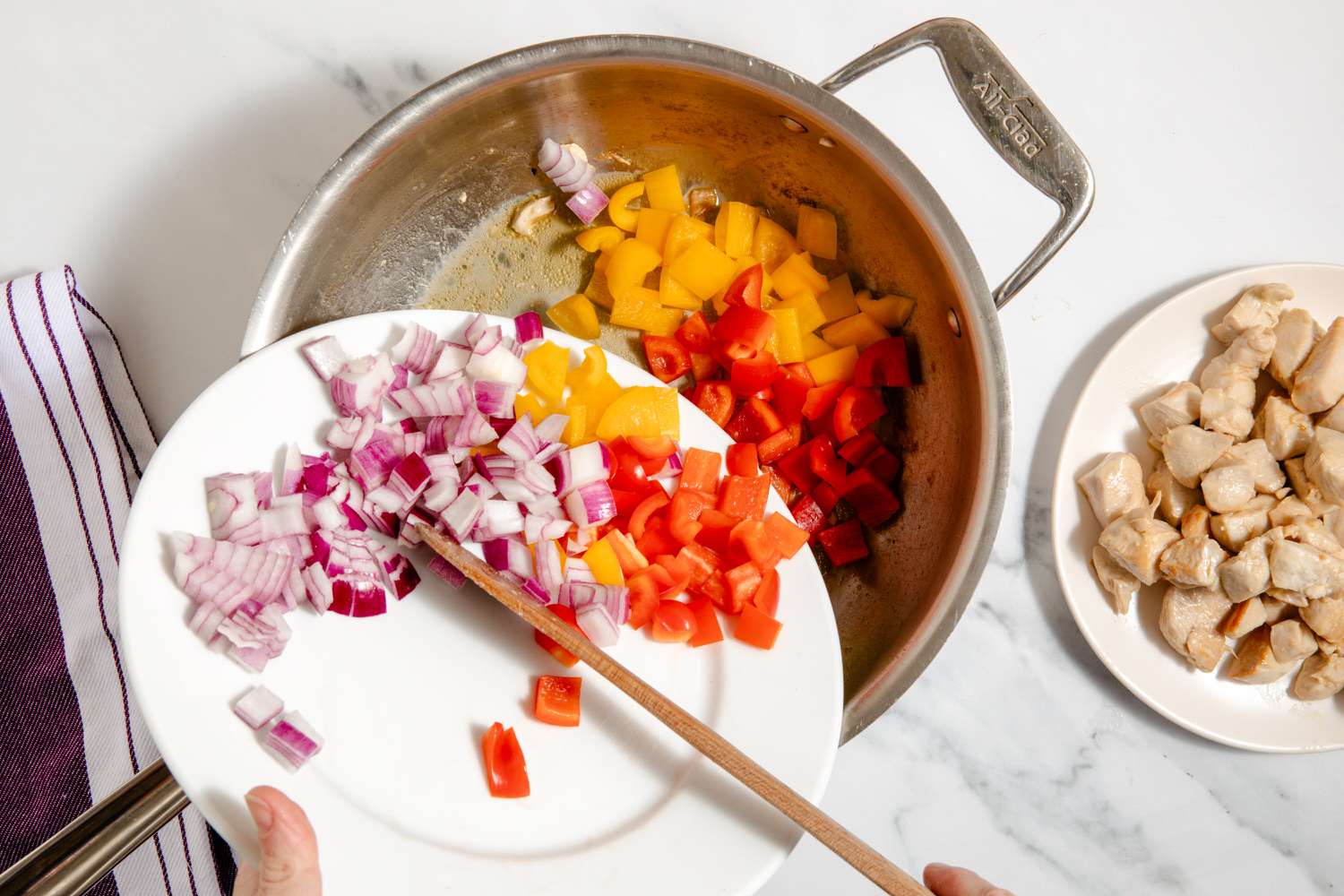 overhead view of hands pouring in chopped onions and peppers into pan, cooked chicken on a plate on the side for One-Pan Chicken Fajita Pasta recipe