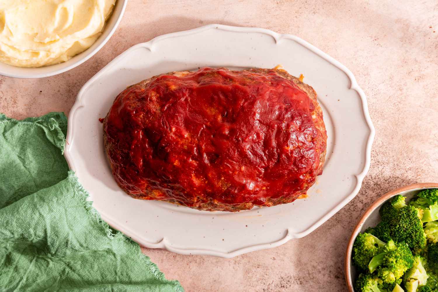 Easy Meatloaf on a Platter and Surrounded by Two Bowls of Sides (One Mashed Potatoes and Another Broccoli) and a Sage Kitchen Towel