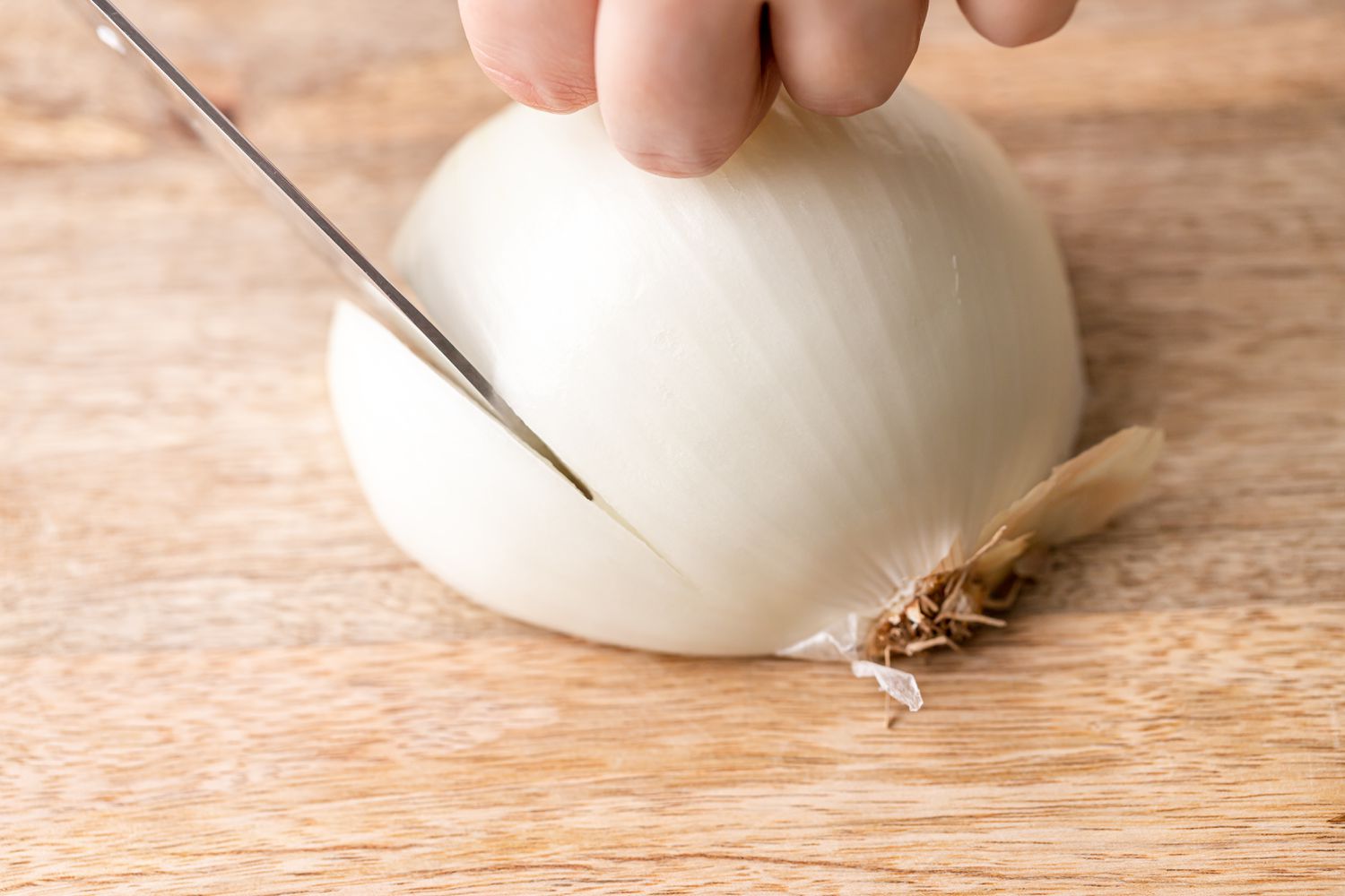 A halved white onion, resting on a cutting board, being sliced with a chef's knife.