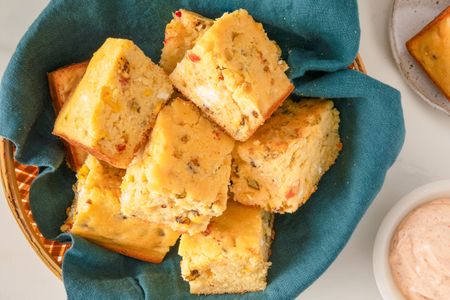 A basket of sliced cornbread on a blue cloth accompanied by a dish of sauce