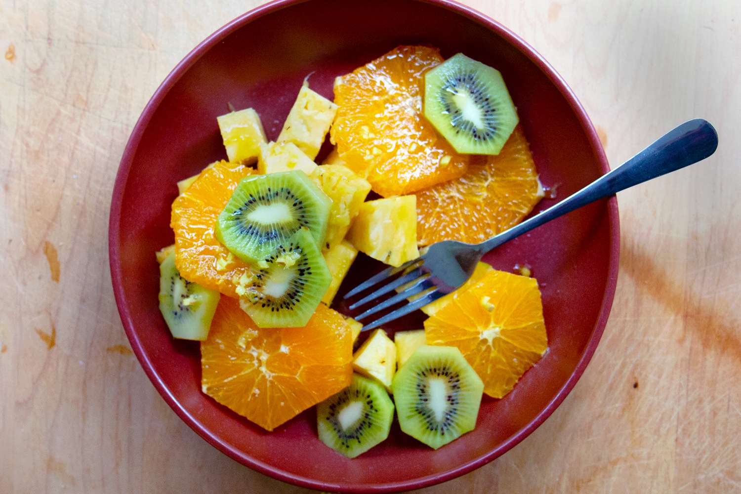 A red bowl with sliced oranges, kiwi, and pineapple, with a fork placed inside the bowl