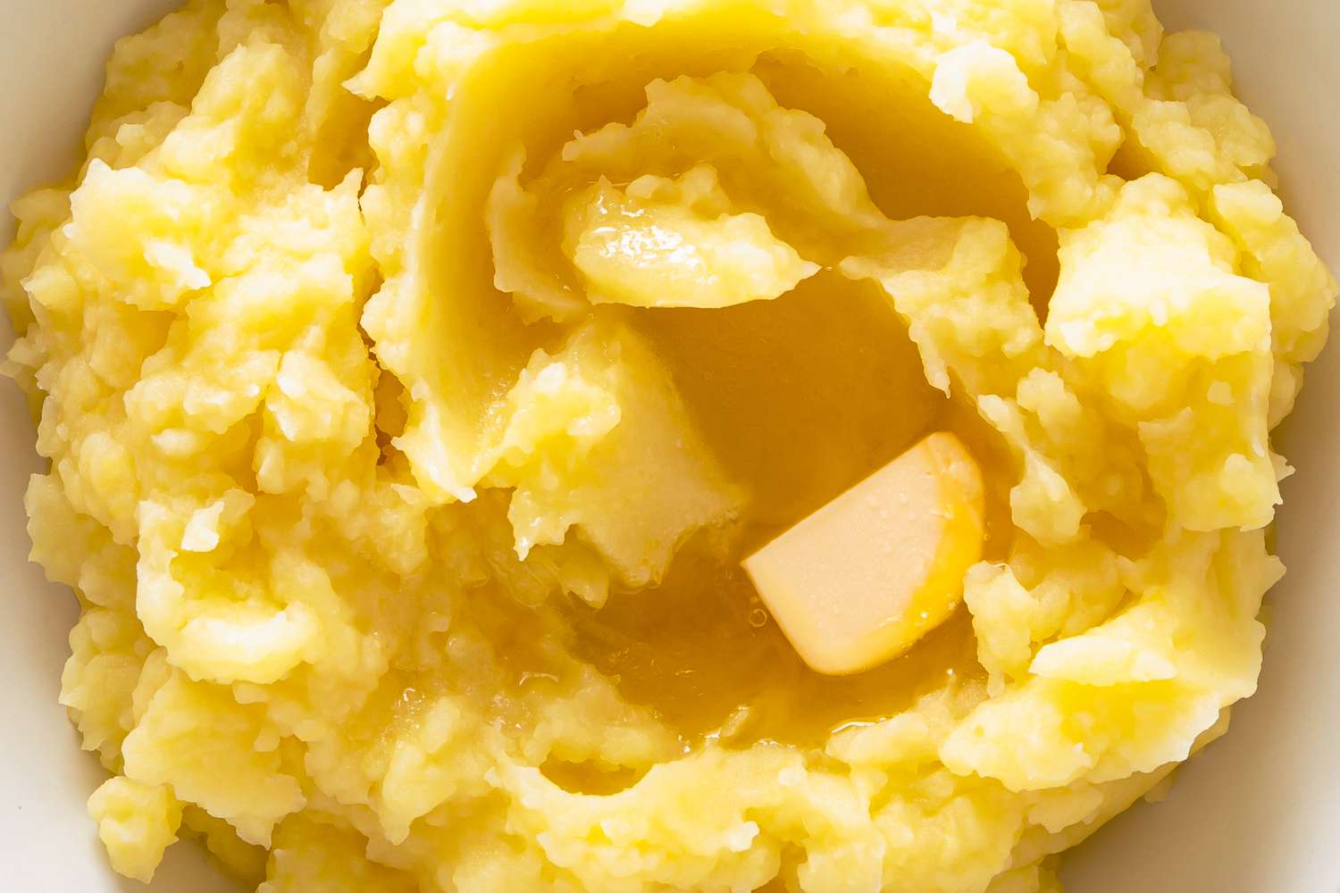 Closeup overhead view of a white bowl of creamy mashed potatoes with a pat of butter melting in the middle