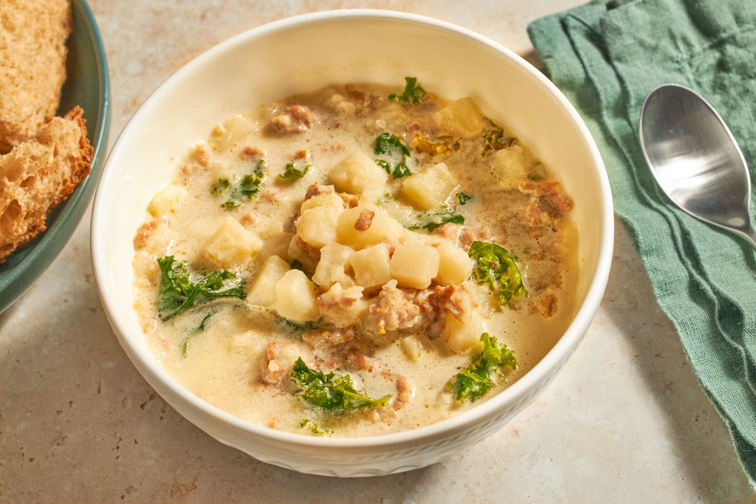 A bowl of sausage kale and potato soup with a slice of bread and a spoon nearby