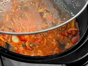 A slow cooker with a tomatobased stew visible through a glass lid containing meat onions and other vegetables