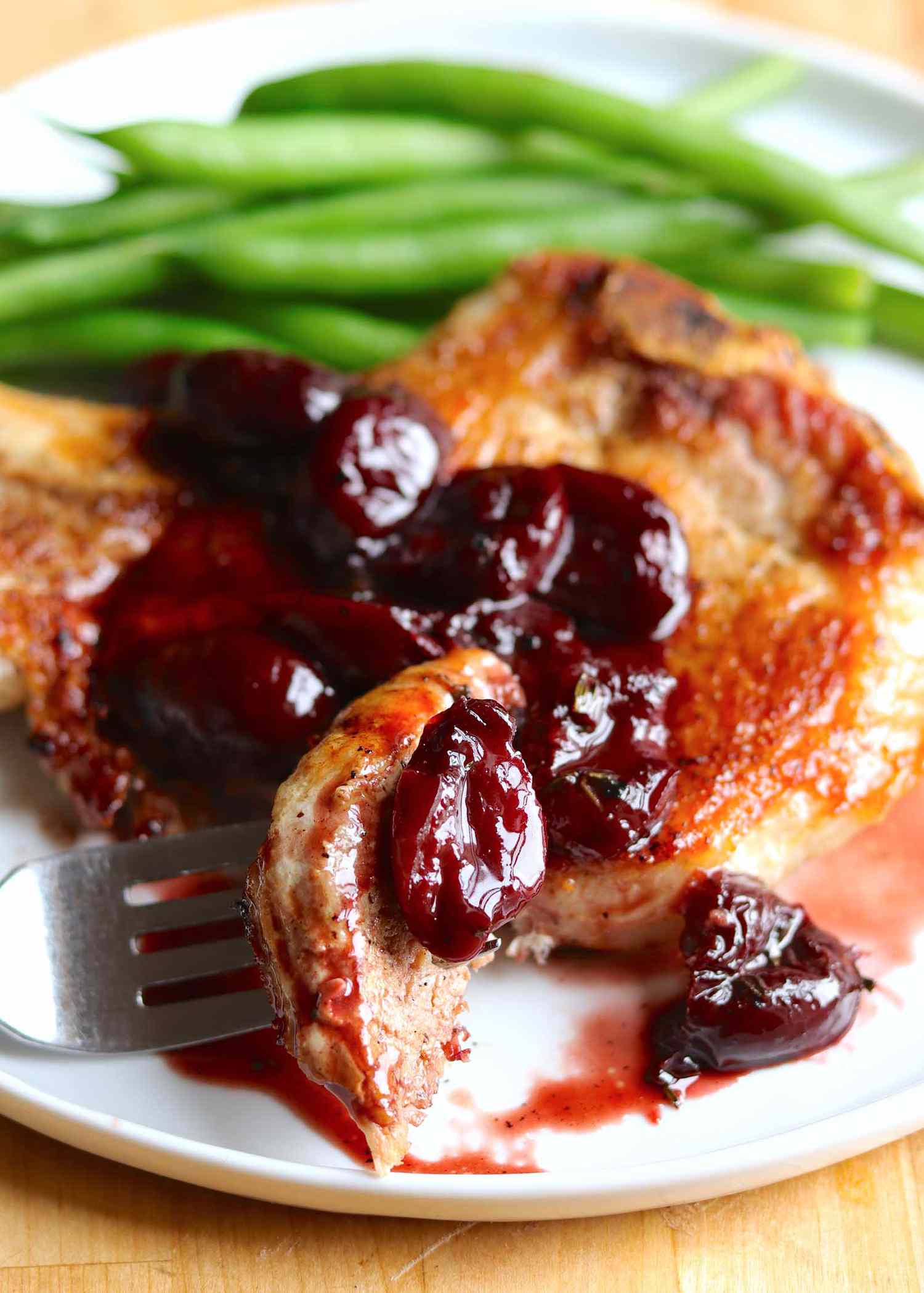 Close up of the best pork chop on a plate topped with a cherry sauce. A forkful of pork chop and cherry are in front.