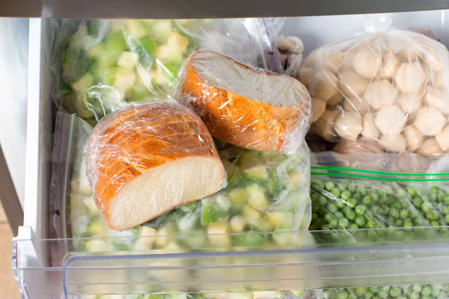 A freezer drawer filled with bread, zucchini, peas, and crackers. 