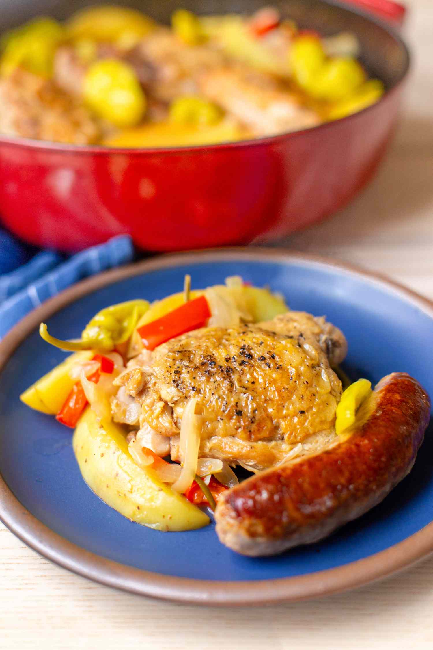 Serving of chicken scarpariello on a plate, and in the surroundings, more in a Dutch oven and blue kitchen towel, all on a wooden countertop