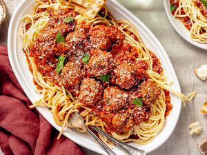 Platter of Spaghetti and Meatballs With a Piece of Toasted Bread and Topped With Fresh Basil Leaves, and in the Surroundings, a Burgundy Table Napkin, a Bowl of Parmesan, Bite Size Bread Pieces Scattered on the Table, and a Plate With More Spaghetti and Meatballs