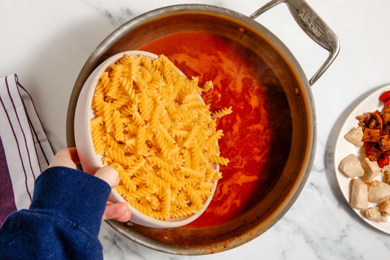 hand pouring rotini into pan with tomato mixture, chicken and veggies on a plate on the side for One-Pan Chicken Fajita Pasta
