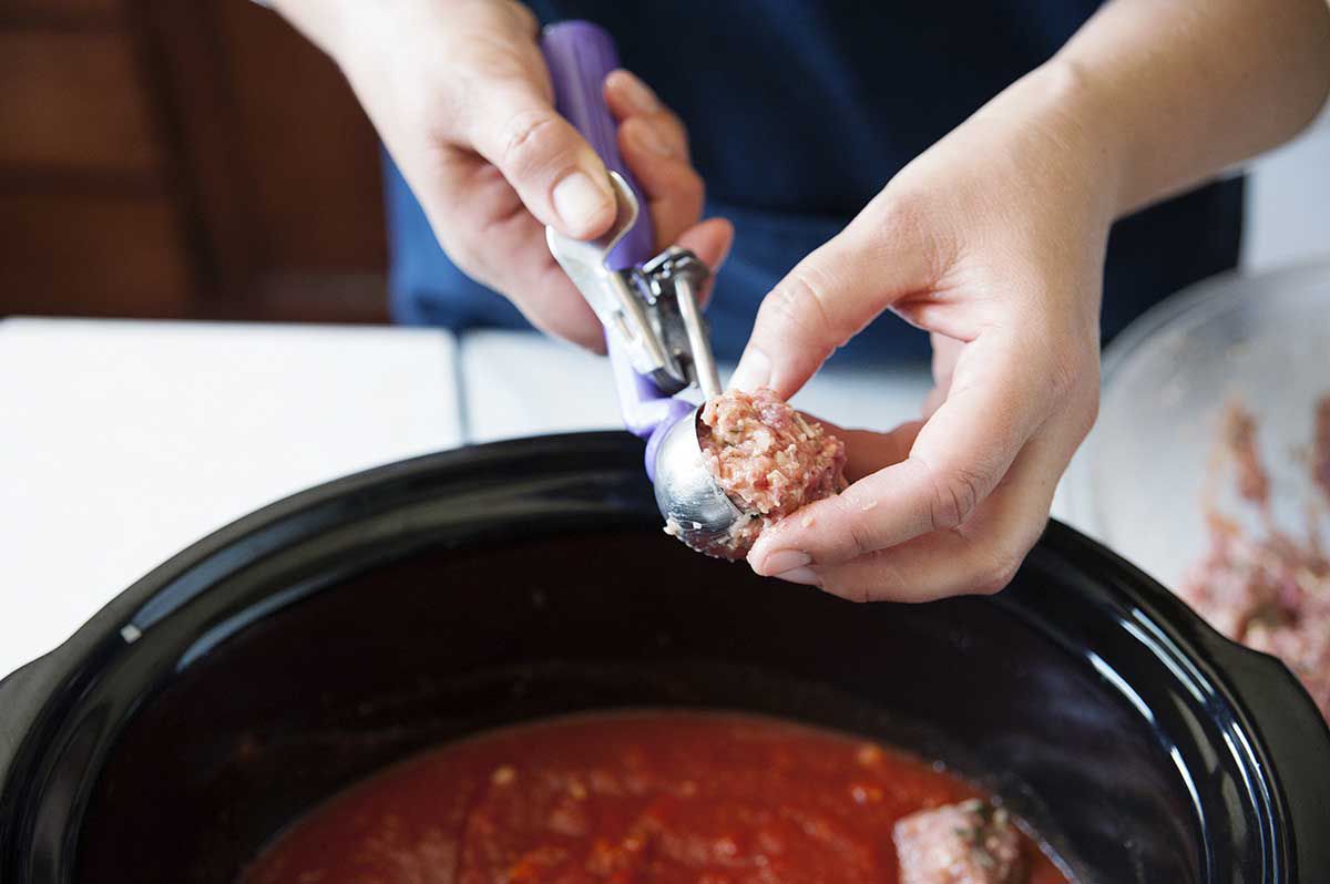 Scooping meatballs with an ice cream scoop and dropping them into a slow cooker filled with tomato sauce.