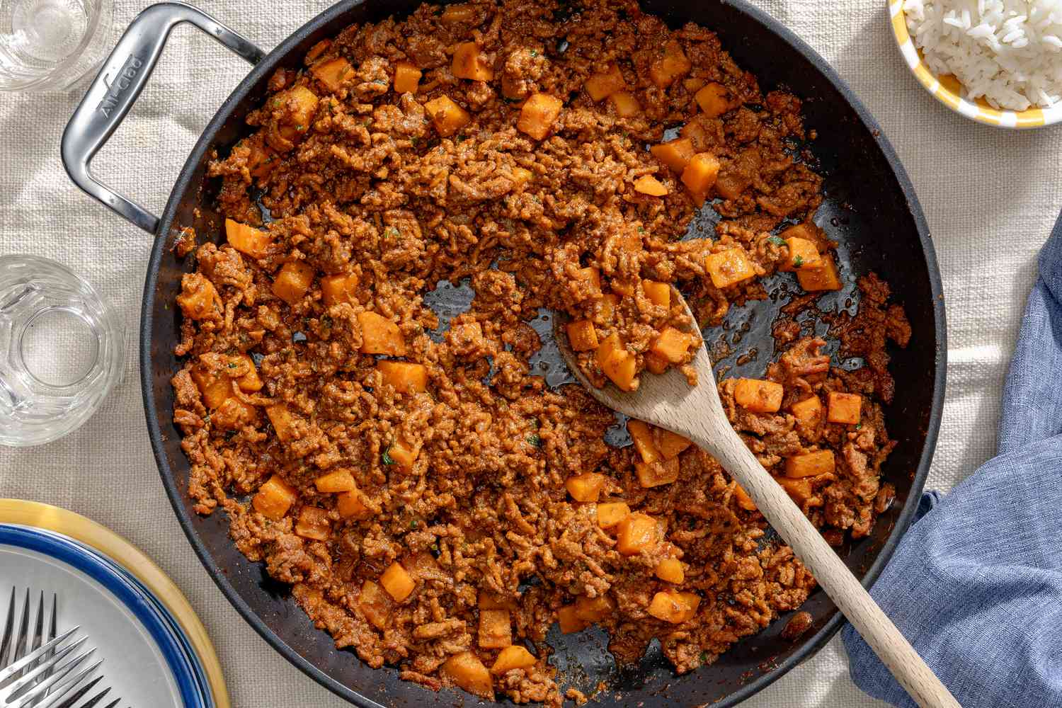 Overhead view of a skillet of picadillo with a wooden serving spoon on a white tablecloth