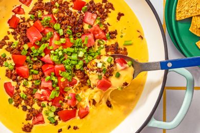 Queso dip with toppings of chopped tomatoes green onions and chorizo served with a spoon and tortilla chips on the side