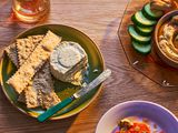 A plate with Boursin cheese and crackers, surrounded by a glass of wine, hummus and cucumbers