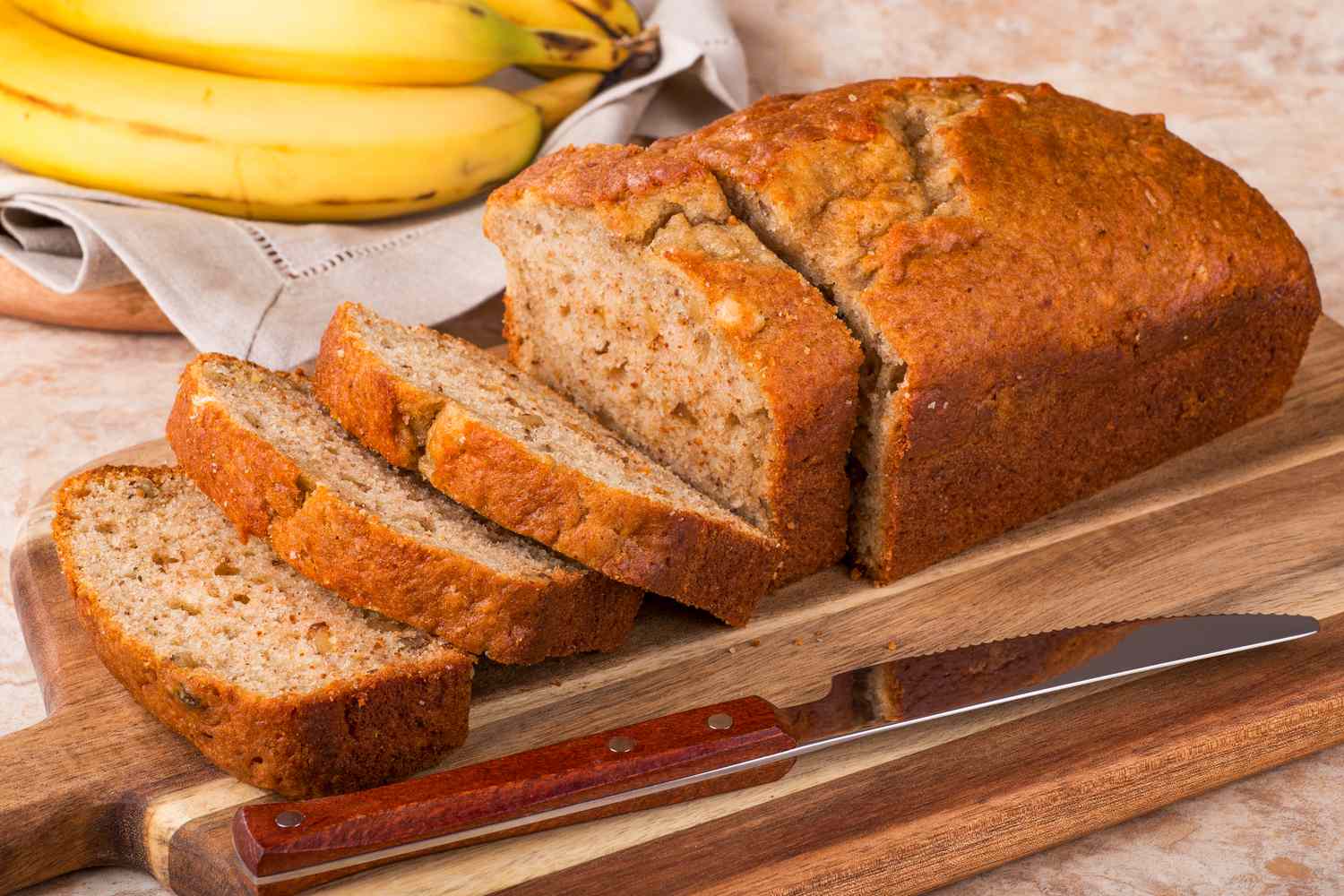A partly sliced banana bread on a cutting board, with bananas in the background