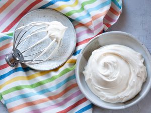Whipped cream frosting with cream cheese in a bowl with a whisk next to it and a striped linen underneath.