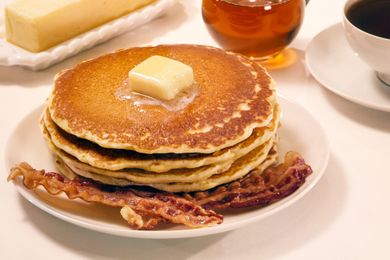 A plate with a stack of pancakes topped with butter accompanied by crispy bacon strips syrup and a coffee cup visible in the background