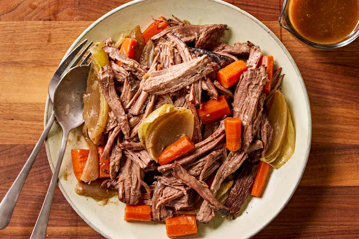 Overhead view of a plate of pot roast with carrots and onions and a fork and spoon all next to a cup of gravy on a wooden tabletop