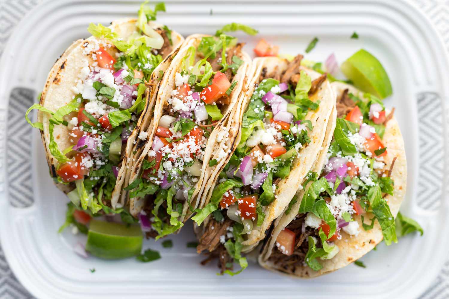 Four tacos filled with shredded pork, lettuce, diced tomatoes, crumbled cheese, and onions on a plate