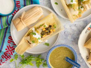 Poblano, Corn, and Cheese Tamales Topped with Salsa Verde, Crema, and Cilantro on Two Plates and Surrounded by a Bowl of Salsa Verde, a Bowl of Crema, and a Platter with More Tamales All on a Table Runner