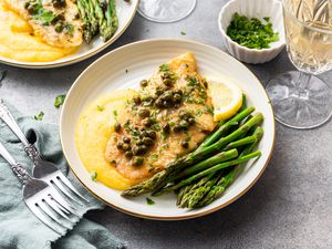 Sole Piccata with Asparagus, Polenta, and Lemon Slice Topped with Capers Surrounded by Another Plate of Sole Piccata, Forks, a Glass, and a Bowl of Herbs 