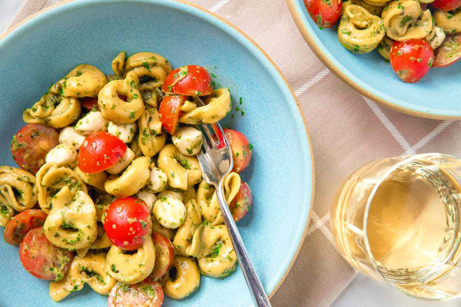 A tortellini pesto salad with halved red cherry tomatoes and tiny mozzarella balls; the salad is in blue bowls on a table with a glass of white wine off to the side