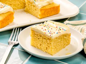 A slice of vanilla cake with frosting and sprinkles on a white plate, next to a fork and a napkin, with a tray of cake pieces in the background