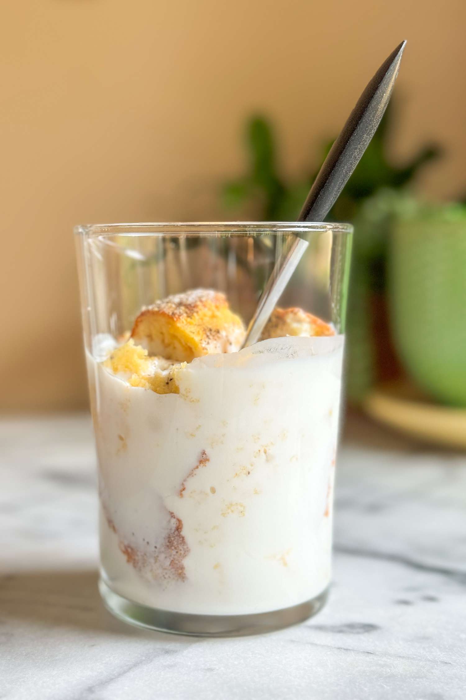 Side view of a glass of cornbread and buttermilk and a spoon resting on a marble countertop with green dishes and a plant in the background