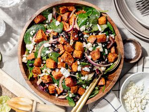 Bowl of Roasted Sweet Potato Salad Topped with Crumbled Feta Cheese, Surrounded by Stack of Plate with Forks, a Small Bowl of More Feta Cheese, a Small Bowl of Salt, a Cutting Board with Lime Wedges, a Kitchen Towel, and Empty Glasses