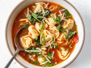 A serving of tortellini soup with a tomato based broth in a bowl, topped with parmesan cheese, with a spoon in the bowl