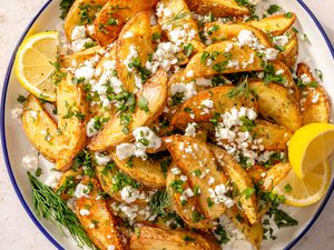 Overhead shot of a serving plate with Greek-style baked potato wedges, garnished with crumbled feta, dill and lemon wedges