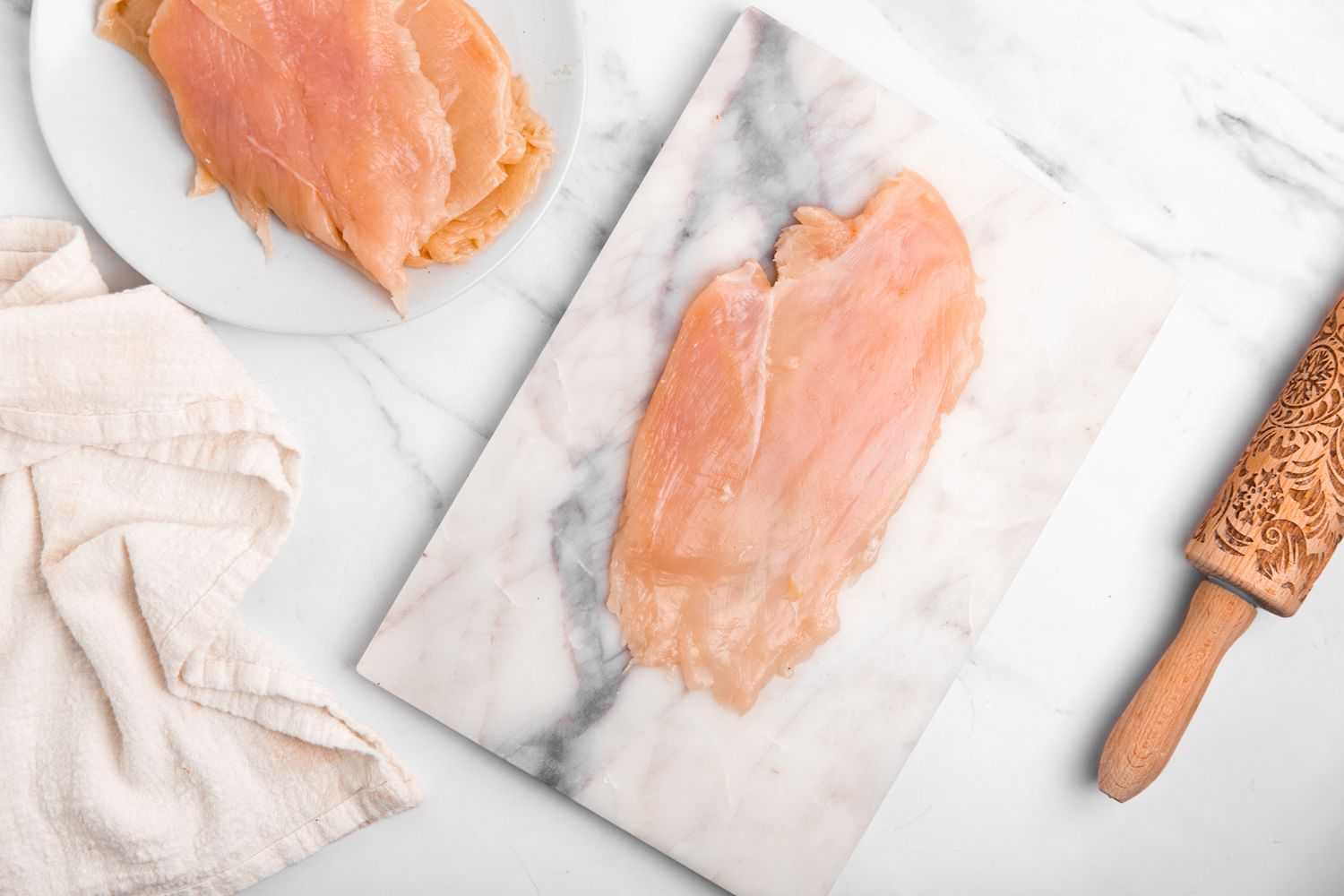 Thinly Sliced Chicken Breast on a Marble Slab Next to a Plate With More Thinly Sliced Chicken Breast, a White Kitchen Towel, and a Rolling Pin