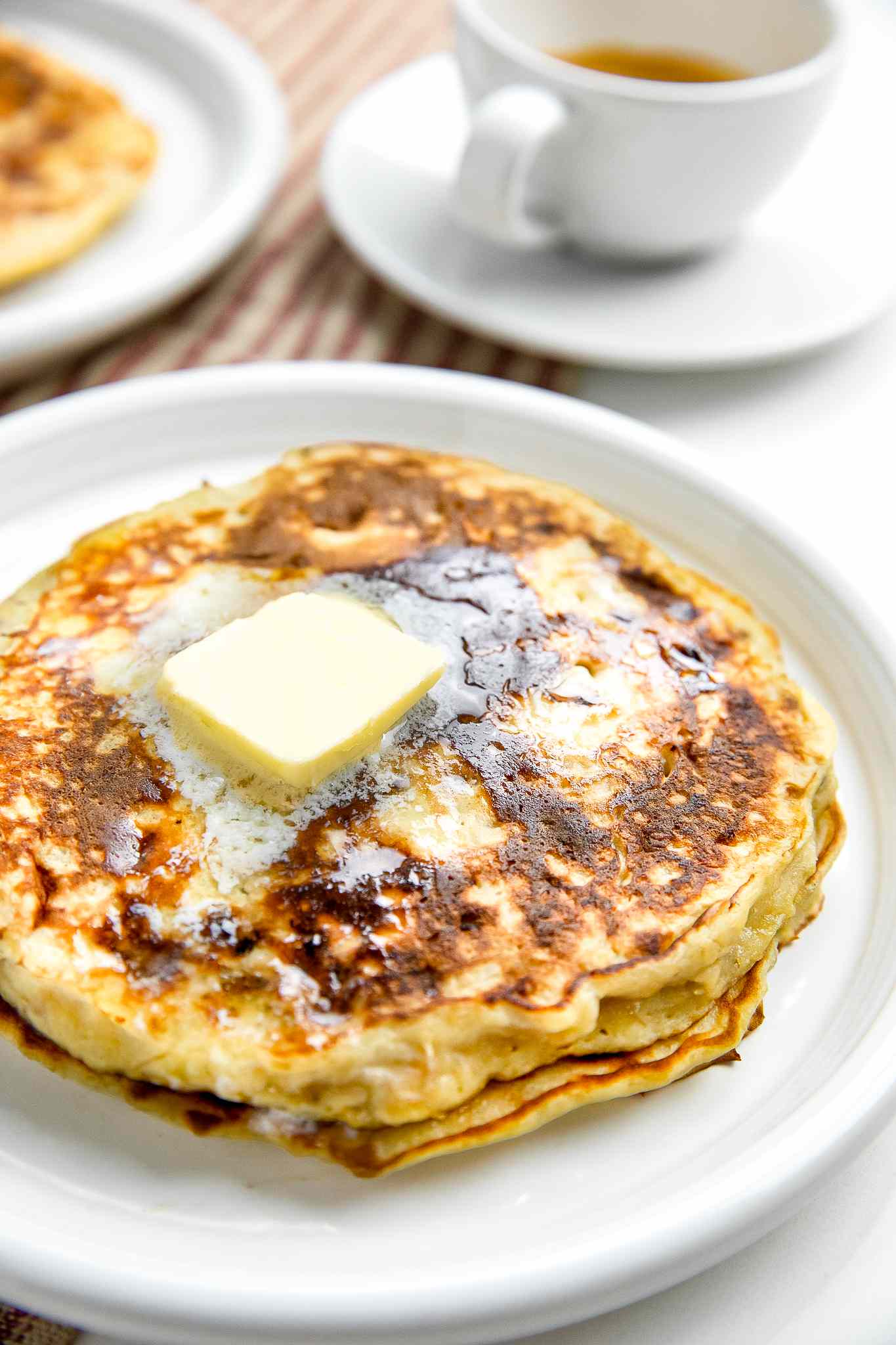 Plate with a Stack of 3-Ingredient Banana Pancakes with Butter, Surrounded by a Plate with More Pancakes and a Cup of Coffee