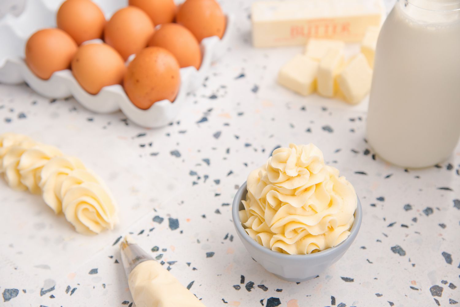 The BEST German buttercream piped in a bowl with eggs, butter, and cream behind it.