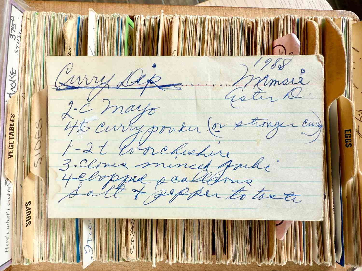 A handwritten recipe card for curry dip placed over a wooden recipe box containing categorized dividers